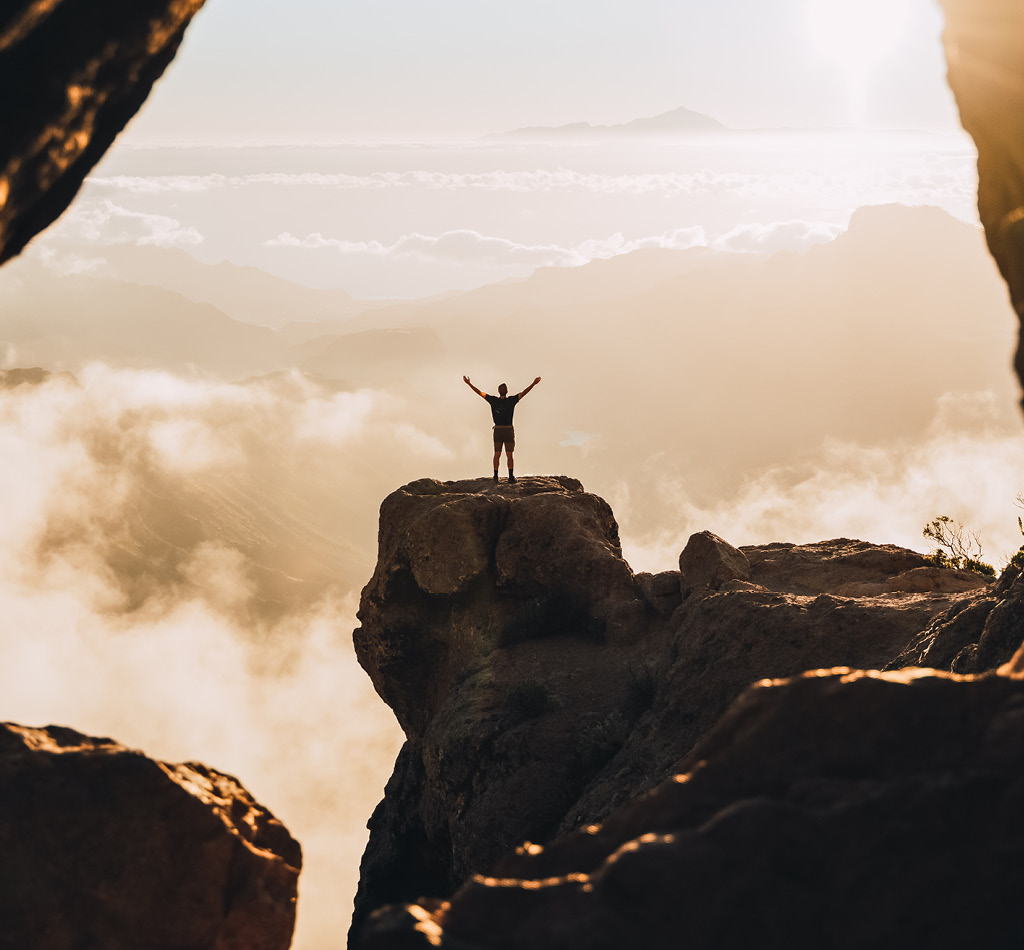 Man standing on the top of a high cliff during the sunset with raised hands.